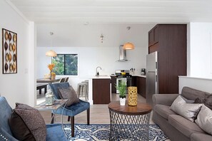 A modern living room with a sofa, a coffee table, and a dining area with a table and chairs. There is a kitchen area with a stove, refrigerator, and cabinets. - Fabulous Townhouse, close to beach, town and harbor (Dana Point)