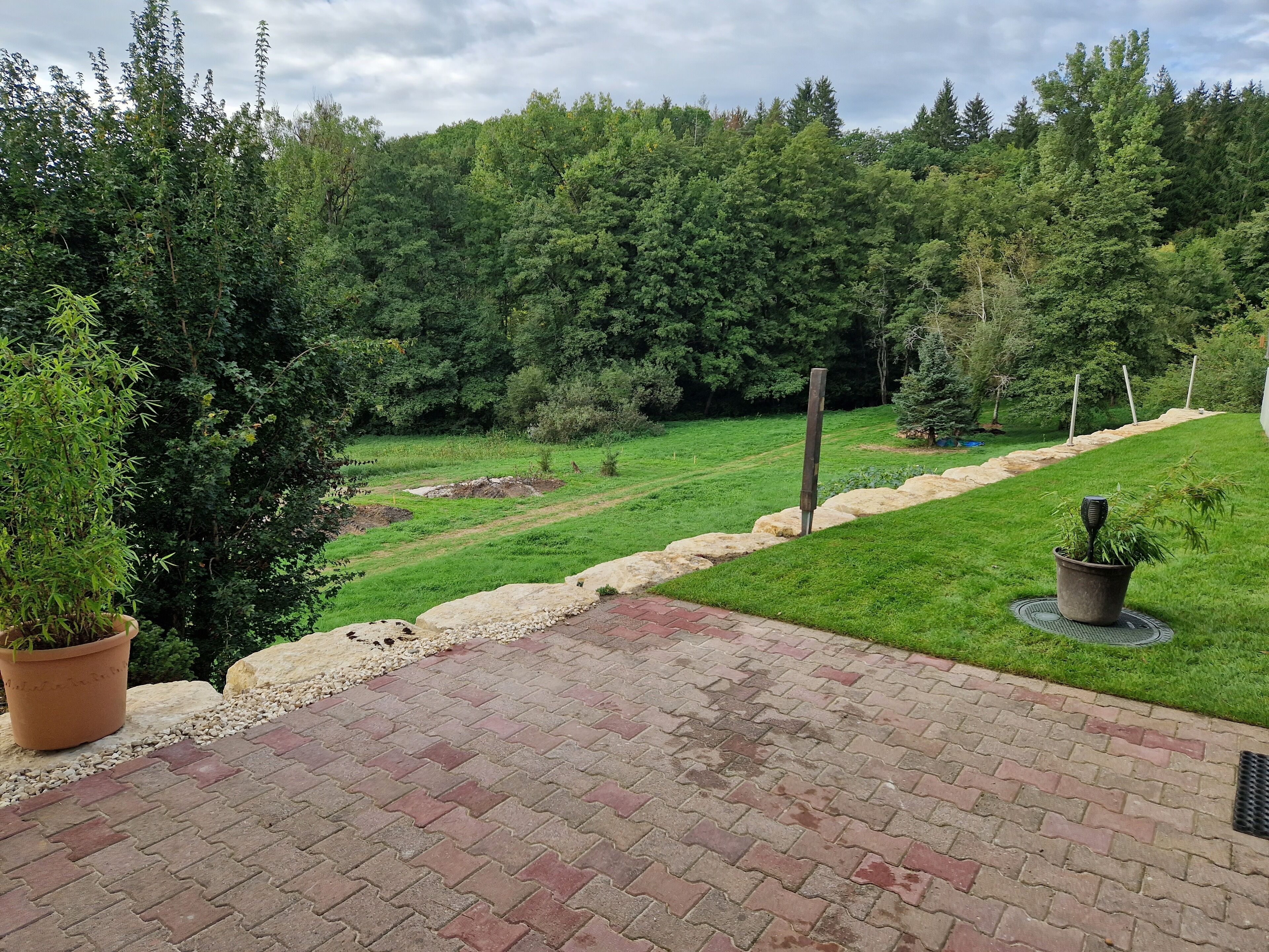 Eine gepflasterte Terrasse mit Ziegelmuster, umgeben von einer Rasenfläche mit Bäumen und einer Steingrenze.