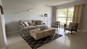 A spacious living room with a beige sofa, a coffee table, and two armchairs. A large window provides natural light, and a 'RELAX' sign hangs on the wall.