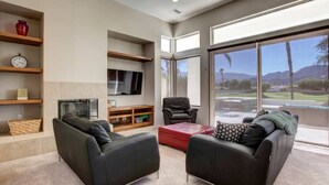 A modern living room with two black leather sofas, a red ottoman, a flat-screen TV, and a fireplace. Large windows offer a view of a landscaped area and mountains. - Waterslide, heated pool home, PGA West 6th tee box (La Quinta)