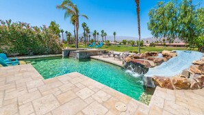 A swimming pool with clear water, surrounded by palm trees and a waterfall feature. - Waterslide, heated pool home, PGA West 6th tee box (La Quinta)