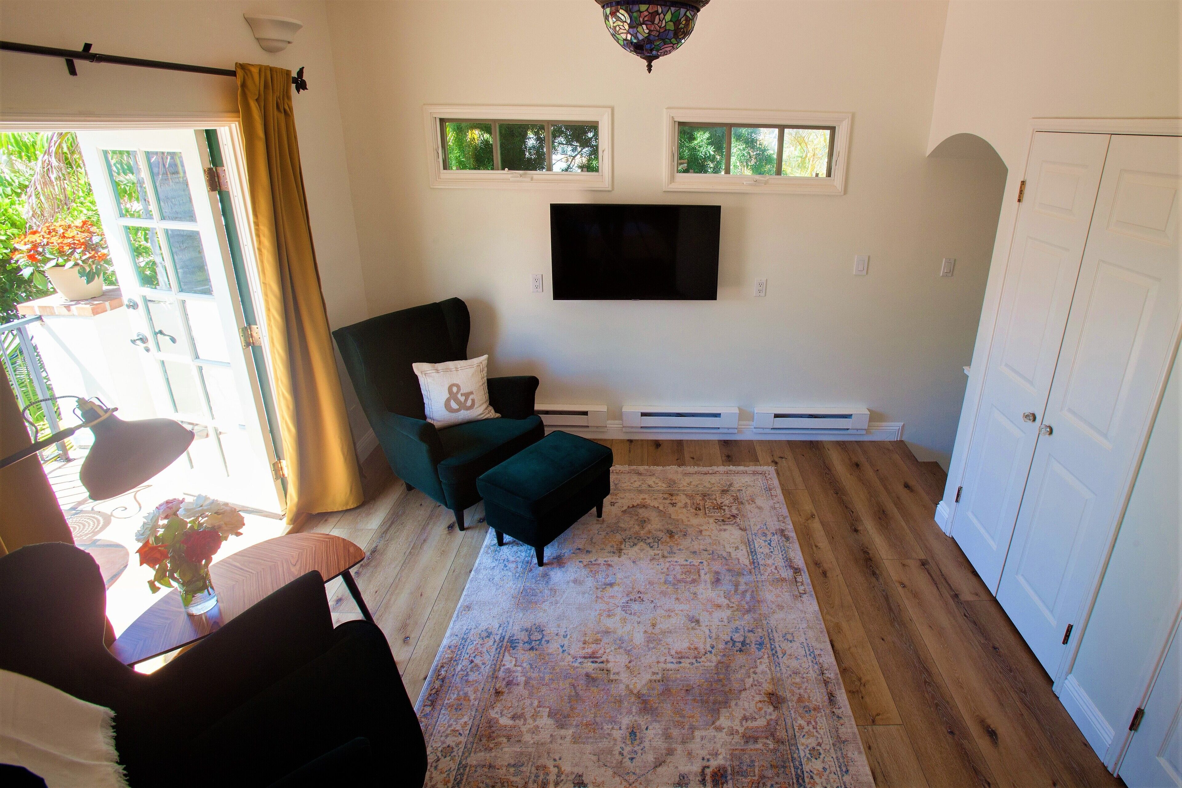 A living room with a sofa, ottoman, and a large rug. There is a TV mounted on the wall, a window with a view of greenery, and a door to the right.