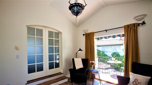 A room with a large window, a chair, a small table with a lamp, and a sofa with a cushion. - Garden Cottage Casita (San Clemente)