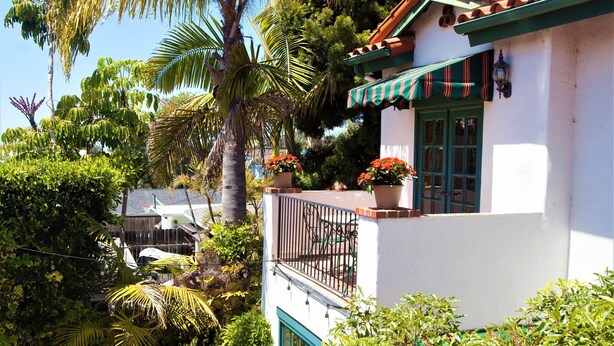 A white building with a red-tiled roof, a balcony with potted plants, and a stone pathway leading to a patio area. - Garden Cottage Casita (San Clemente)