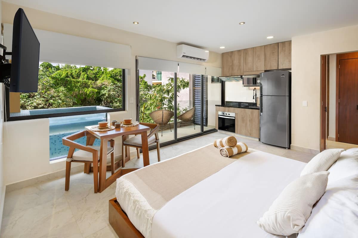 A modern hotel room with a balcony, a dining area, a kitchen, and a large bed.