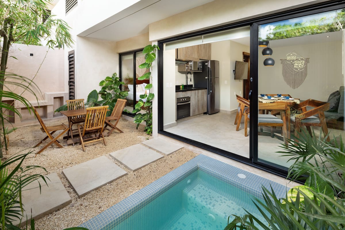 An outdoor patio with a pool, a dining area with wooden furniture, and a kitchen visible through glass doors.