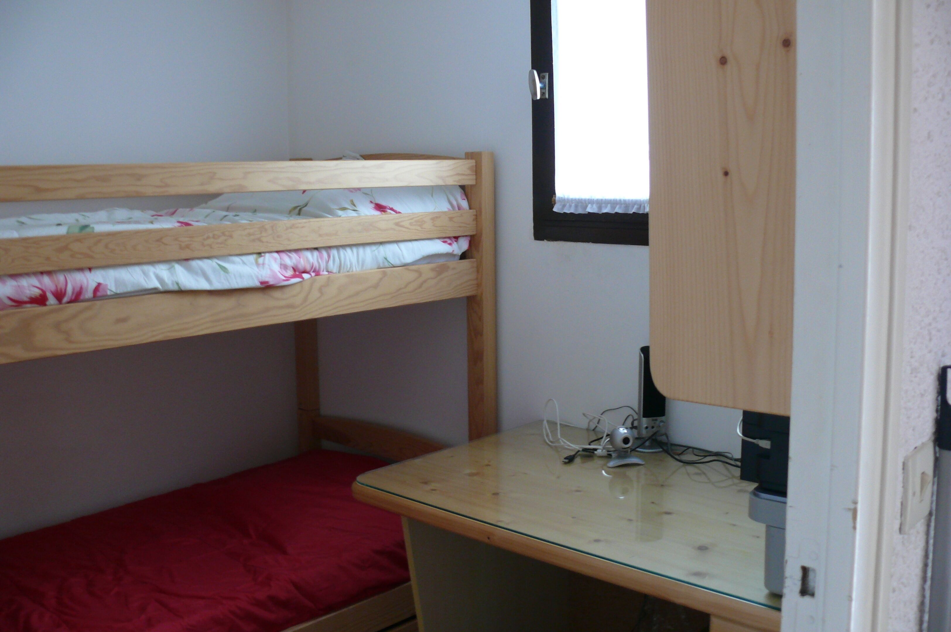 A bunk bed with a wooden frame and a single bed below. There is a small desk with a computer and a window above the desk.