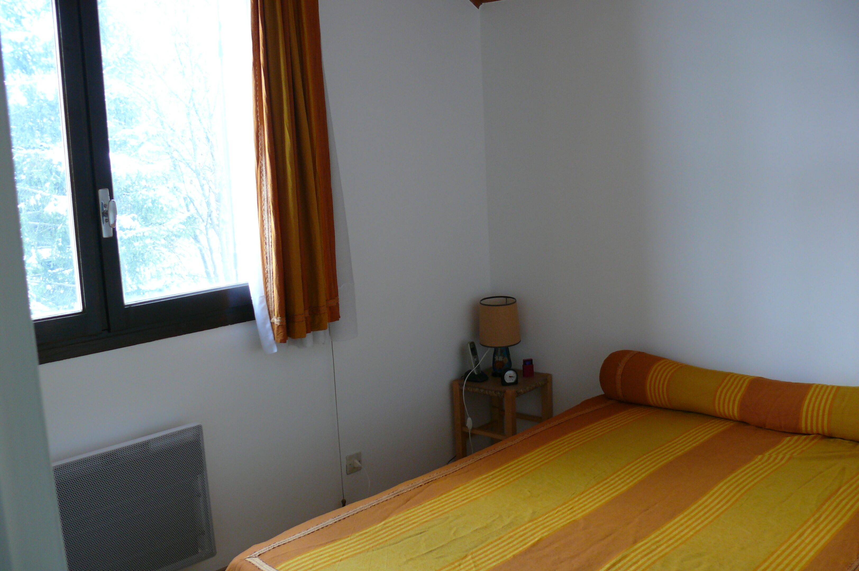Une chambre à coucher avec un lit, une fenêtre avec des rideaux, une table de chevet avec une lampe et un radiateur.