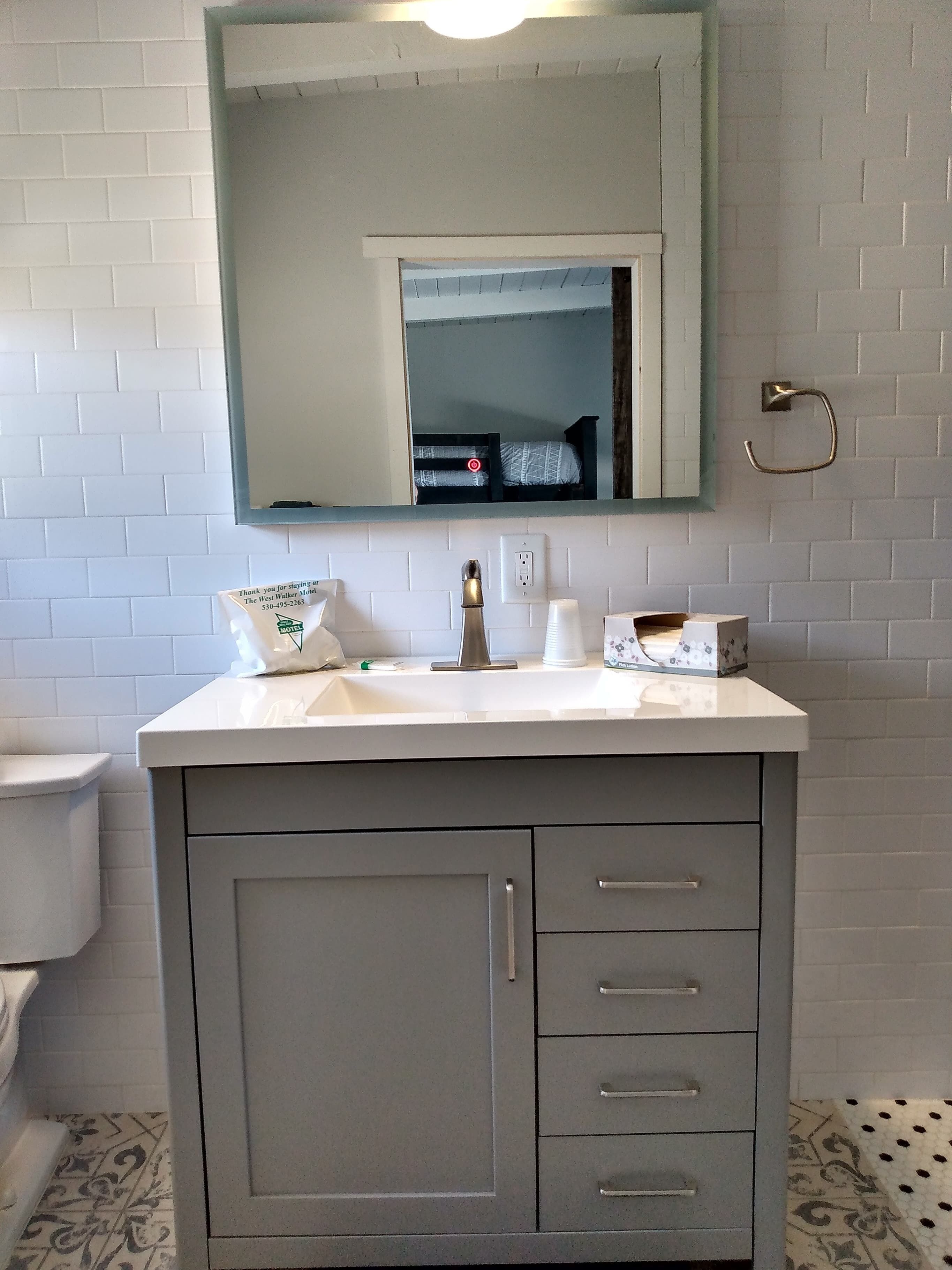 A bathroom with a white sink, a mirror, and a toilet. There is a sign on the sink and a towel dispenser.