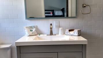 A bathroom with a white sink, a mirror, and a toilet. There is a sign on the sink and a towel dispenser.