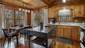 A wooden cabin interior with a dining table, chairs, a kitchen with wooden cabinets, and large windows offering a view of trees. - Welcome to Bearadise! Charming cabin 10 minutes to downtown Blue Ridge! (Cherry Log)