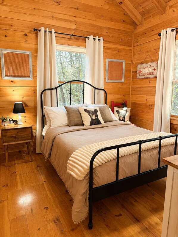 A wooden cabin bedroom with a bed, nightstand, lamp, and a sign indicating a flea market. - Welcome to Bearadise! Charming cabin 10 minutes to downtown Blue Ridge! (Cherry Log)