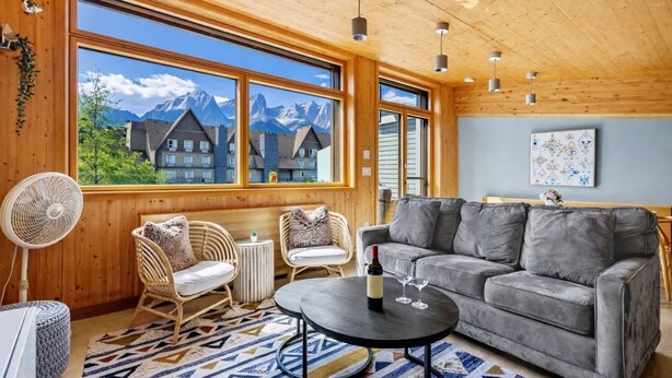 A cozy living room with a grey sofa, wicker chairs, a small round table, and a fan. Large windows offer a view of mountains and buildings. - Dolomites #203 - LEED Gold Certified! (Canmore)