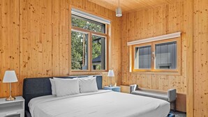 A bedroom with a large bed, two bedside tables, a chair, and a window with a view of trees. - Dolomites #203 - LEED Gold Certified! (Canmore)