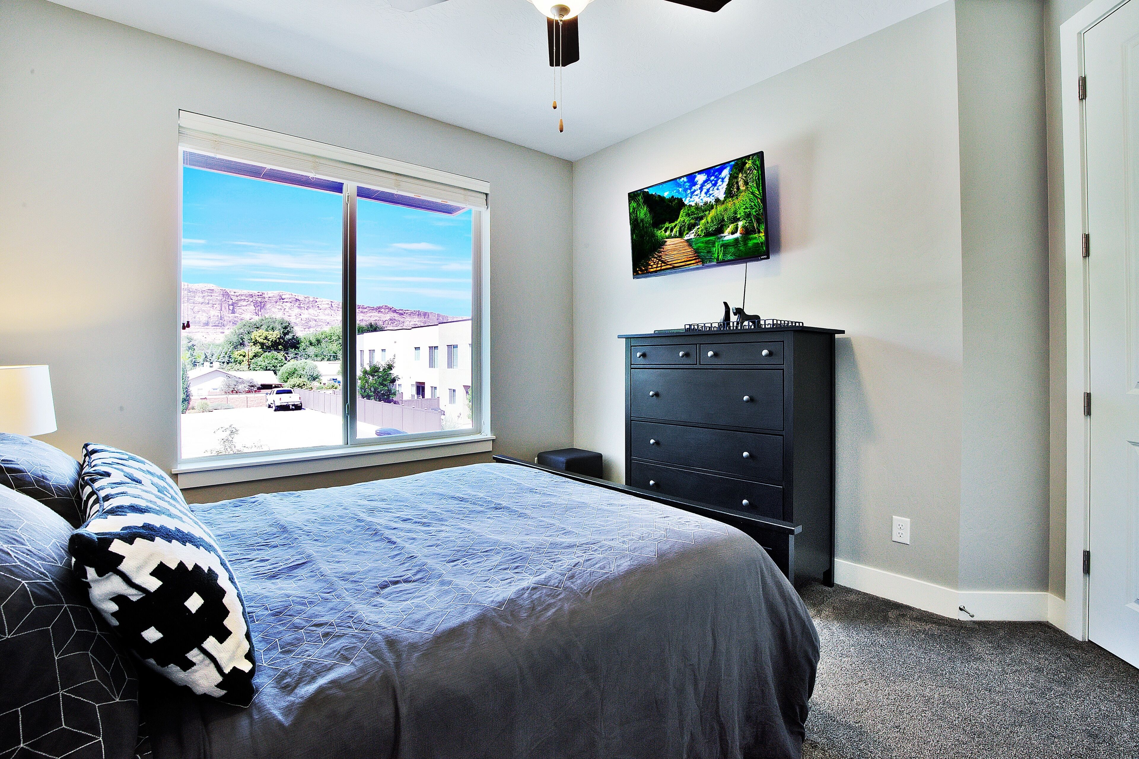A bedroom with a large window, a bed with patterned pillows, a dresser, and a mounted TV.