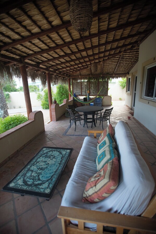 A covered patio with a thatched roof, a round table with chairs, a sofa with cushions, and a patterned rug.