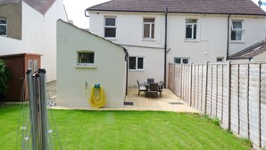 Un jardin à l’arrière d’une maison, comprenant une terrasse, une pelouse et une maison dotée de nombreuses fenêtres et d’un toit recouvert de tuiles.