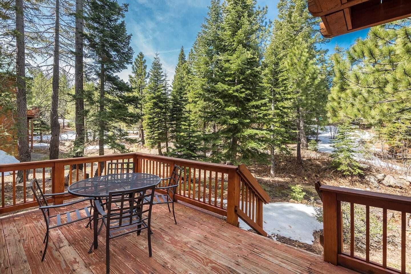 A wooden deck with a round table and chairs, surrounded by tall pine trees and a view of a snowy landscape.