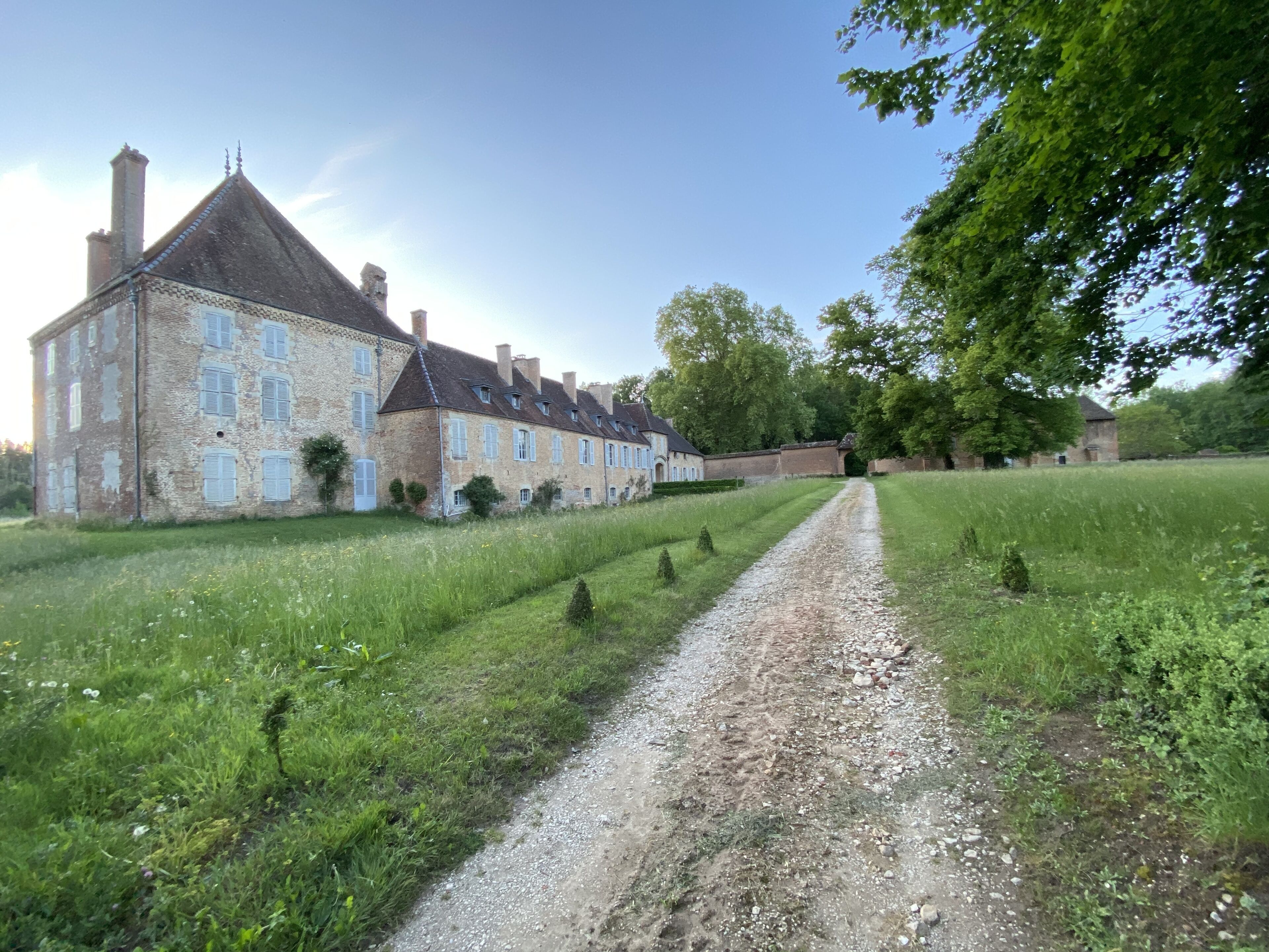 Ein großes, historisches Steinhaus mit vielen Fenstern und einem markanten Schornstein, umgeben von einem gepflegten Rasen und einem Kiesweg, der zu einem Tor führt.