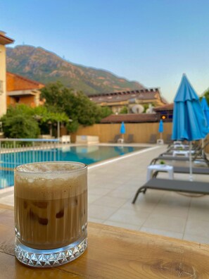 Un vaso de café helado sobre una mesa de madera junto a una piscina con sillones de playa y sombrillas, con vistas a una montaña y un edificio con techo de tejas.