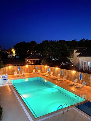 Una piscina rectangular con solárium empinado, rodeada de sillones de playa y sombrillas, y un pequeño bar junto a la piscina.