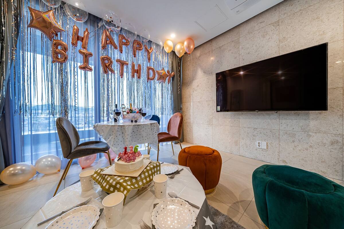 A modern room with a round table set for a birthday celebration, featuring a cake with candles, a TV mounted on the wall, and a large window with curtains.