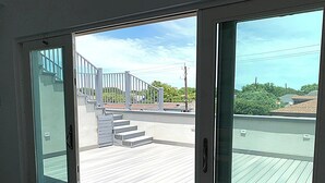 A view through a sliding glass door to a wooden deck with a metal railing and a staircase leading to a rooftop.