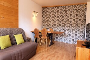 Une pièce chaleureuse avec un plancher en bois, un canapé marron agrémenté de coussins verts, une table à manger en bois avec des chaises, et un mur orné d’un motif.