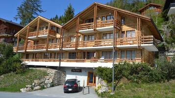 Un chalet en bois à plusieurs étages, doté de balcons, d’une entrée surmontée d’un mur en pierre et d’une voiture garée devant.