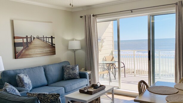 Sala de estar com sofá azul, mesa de centro e mesa de jantar com cadeiras. Há uma janela grande com vista para a praia e oceano.