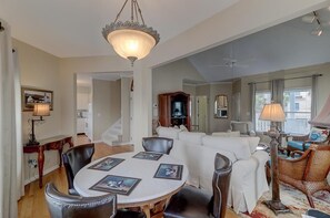 A spacious living area with a round dining table, leather chairs, a white sofa, and a wicker armchair. A chandelier hangs from the ceiling, and there is a television in the background.