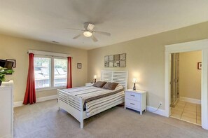 A bedroom with a bed, bedside tables, a ceiling fan, a window with curtains, and a door leading to a shower area. - 26 42nd Avenue (Isle of Palms)