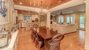 A spacious dining area with a long wooden table and wicker chairs, a kitchen with white cabinets, and a living area with a sofa and large windows. - 2602 Hartnett Boulevard (Isle of Palms)