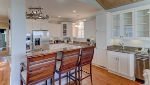 A modern kitchen with white cabinetry, a central island with bar stools, stainless steel appliances, and a wooden ceiling. - 21 Ocean Point (Isle of Palms)