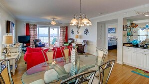 A spacious living area with a glass dining table, red sofas, and a modern chandelier. - 1010 Ocean Palms 201 (Isle of Palms)