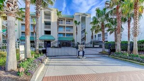 A gated entrance to a multi-story hotel with a green roof, palm trees, and a paved walkway. - 1010 Ocean Palms 201 (Isle of Palms)