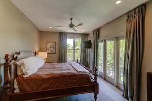 A bedroom with a large bed, a ceiling fan, a television, and a balcony with curtains.