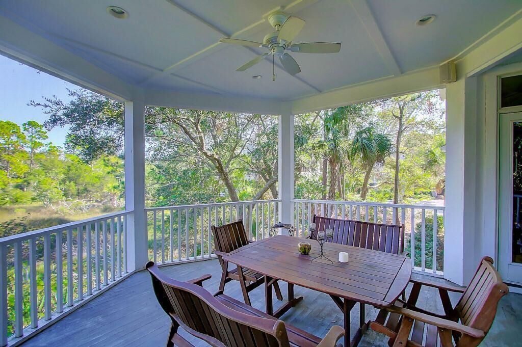 A covered outdoor patio with a table and chairs, surrounded by white railings, overlooking a natural landscape with trees and a body of water.