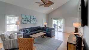 A spacious living room with a large sectional sofa, a coffee table, and a television. There are two windows with blinds, a ceiling fan, and a door leading to an outdoor area. - 903 Ocean Boulevard (Isle of Palms)