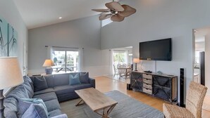 A spacious living area with a sofa, coffee table, and a television. There is a ceiling fan, a dining area with chairs, and a view of the outdoors through large windows. - 903 Ocean Boulevard (Isle of Palms)