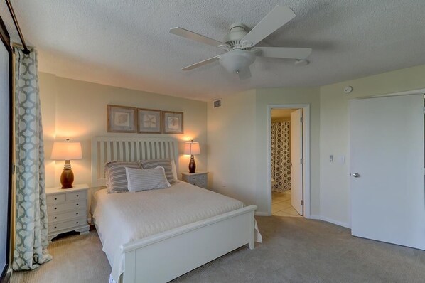 A bedroom with a white bed, two bedside tables with lamps, a ceiling fan, and a door leading to another room.
