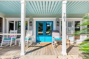 A covered porch with a blue door, numbered 705, and white chairs with teal cushions.
