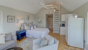 A bedroom with a bed, a nightstand, a desk, and a chair. There is a white ceiling fan, a white refrigerator, and a white oven in the kitchen area. - 8 44th Avenue (Isle of Palms)