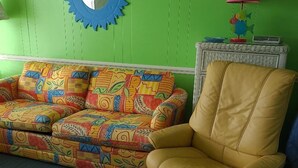 Een kamer met een groene wand, een gestreept bankstel, een gele fauteuil, een ronde spiegel en een klein tafeltje met een lamp.