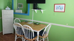 Een kamer met een groene muur, een houten tafel met vier witte stoelen, een witte dressoir, een flatscreen televisie aan de muur bevestigd, en een lijst met een foto erboven.