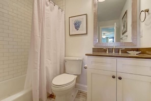Un baño con inodoro blanco, cortina de ducha, un cuadro enmarcado en la pared y un espejo grande sobre un lavabo con encimera.