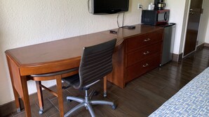 A hotel room with a wooden desk, a black office chair, a television mounted on the wall, a coffee maker, and a microwave on a cabinet. - RW Hotel (Kissimmee)