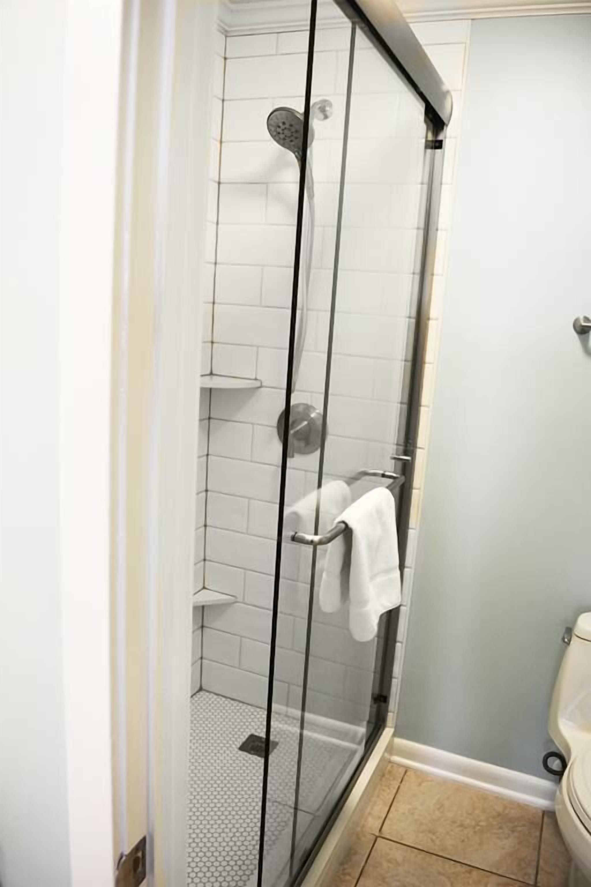 A glass-enclosed shower with a white tiled wall and a white toilet in a bathroom.