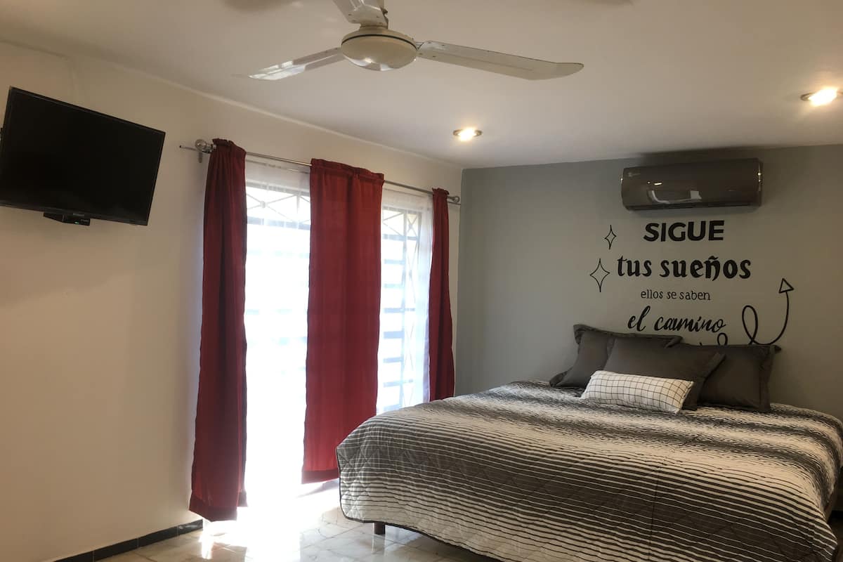 A bedroom with a bed, a ceiling fan, a flat-screen TV, red curtains, and a motivational quote on the wall.