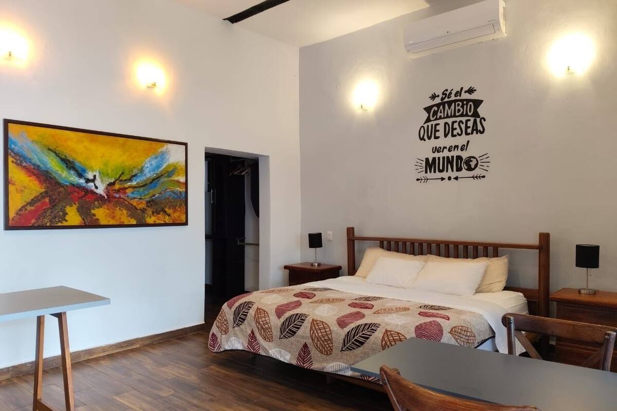 A hotel room with a bed, a desk, a chair, a painting, and a motivational quote on the wall.
